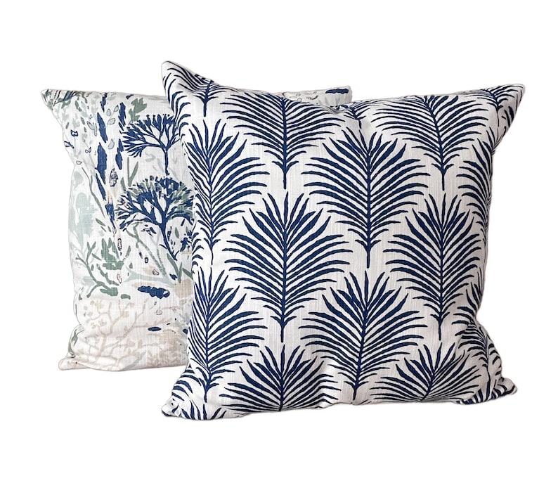 K&ouml;nnte beinhalten: Zwei quadratische Zierkissen. Eines hat einen wei&szlig;en Hintergrund mit einem navyblauen Palmenblattmuster. Das andere hat einen wei&szlig;en Hintergrund mit einem blau-gr&uuml;nen botanischen Design. Ideal f&uuml;r die Dekoration.
