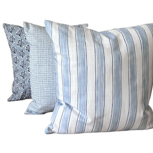 K&ouml;nnte beinhalten: Drei dekorative Kissen in Blau- und Wei&szlig;t&ouml;nen. Die Kissen weisen verschiedene Muster auf: ein korallenartiges Design, ein kleines Gitter und vertikale Streifen. Die Kissen sind in einer Reihe angeordnet, mit dem gestreiften Kissen im Vordergrund.