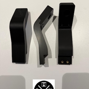 Könnte beinhalten: Drei schwarze, eckige, 3D-gedruckte Komponenten sind auf einer weißen Oberfläche angeordnet. Die Komponenten haben eine matte Oberfläche und unterschiedliche Formen, darunter geschwungene und rechteckige Designs. Ein kreisförmiges Logo mit den Buchstaben K, M und der Zahl 2025 befindet sich unten.