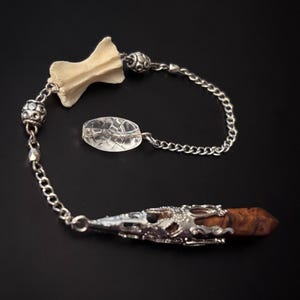 May include: A pendulum featuring a brown, faceted crystal point encased in an ornate silver-toned metal cap, connected by a silver-toned chain to a small, off-white bone fragment and a clear, oval-shaped glass bead with a textured surface.