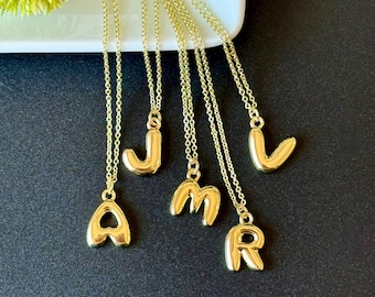 May include: Gold-colored initial necklaces. Each necklace features a single letter pendant, including A, J, M, R, and V, suspended from a delicate chain. The pendants have a rounded, bubble-letter design. The necklaces are displayed against a dark background.