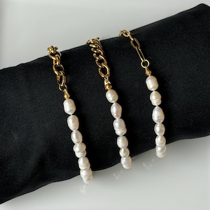 May include: Three gold-toned chain bracelets with dangling white pearl strands. The bracelets are displayed on a black surface, showcasing the contrast between the gold and pearls. The chains have different link styles.