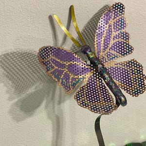 May include: A decorative metal butterfly sculpture with purple and gold wings, featuring a dotted pattern. The butterfly has a slender body and is mounted on a curved metal stand. The sculpture is set against a neutral background.