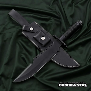May include: A black survival knife with a serrated blade and a black handle, resting in a black leather sheath. The sheath has a belt loop and a small pouch. The text "SCHWARZENEGGER COMMANDO" is visible.