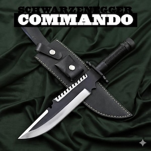 Può includere: Un coltello da commando nero e argento con bordo seghettato, in un fodero di pelle nera. Il testo "SCHWARZENEGGER COMMANDO" è visualizzato sopra il coltello.