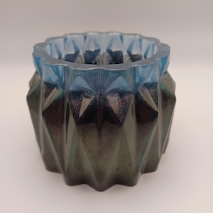 May include: A faceted resin planter in shades of blue and iridescent green. The top rim is a translucent blue, transitioning to a darker, shimmering green base. The geometric design adds a modern touch to this decorative item.