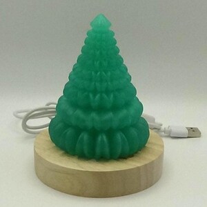 May include: A green Christmas tree-shaped LED light sits on a round wooden base. A white USB cable extends from the base. The tree has a detailed, textured surface and is illuminated. The light is designed for decorative purposes.