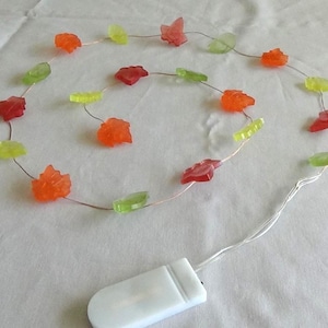 May include: A string of decorative LED lights featuring colorful leaf-shaped ornaments in shades of red, orange, and green. The lights are powered by a small white battery pack, perfect for fall decor.