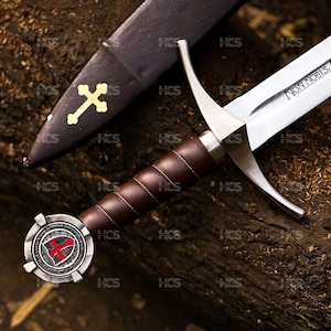 May include: A silver-toned sword with a brown leather-wrapped grip and a circular pommel featuring a red cross emblem. The blade is engraved with the text Non Nobis, Domine. A matching dark scabbard with a gold-colored cross symbol is included.