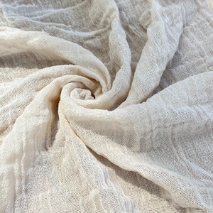 Può includere: Tessuto strutturato bianco sporco con una trama rada, che crea un aspetto morbido e drappeggiato. Il tessuto è disposto in un motivo a spirale, con pieghe e ombre che aggiungono profondità. Il materiale appare leggero e arioso, adatto a vari progetti tessili.