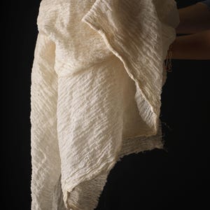 May include: Off-white, textured fabric with a diagonal ribbed pattern. The fabric is draped, revealing its layered construction and frayed edges. The background is a solid black, highlighting the fabric's details. The fabric appears to be lightweight and flowing.