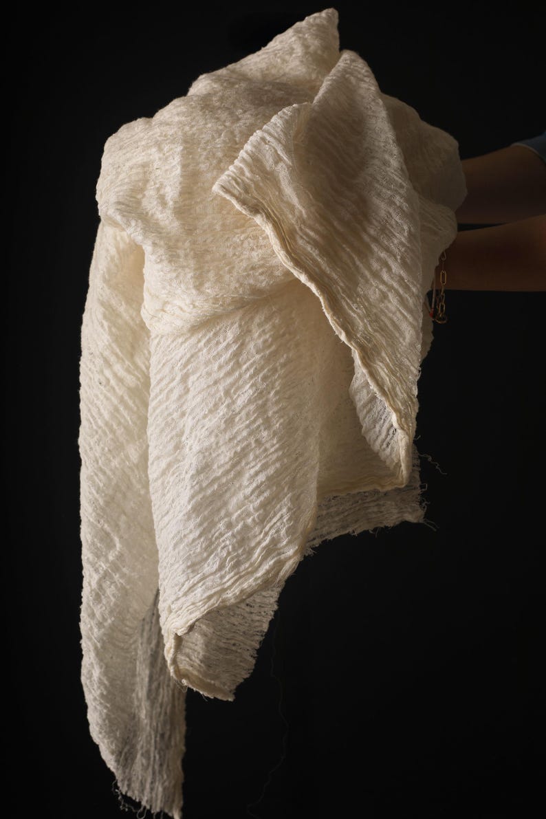May include: Off-white, textured fabric with a diagonal ribbed pattern. The fabric is draped, revealing its layered construction and frayed edges. The background is a solid black, highlighting the fabric's details. The fabric appears to be lightweight and flowing.