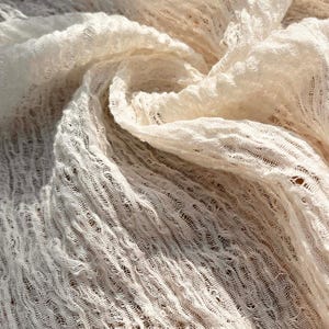 May include: Close-up of a cream-colored, loosely woven fabric. The fabric has a textured, open weave with a slightly wrinkled appearance. The light creates shadows and highlights, emphasizing the texture and drape of the material. The overall effect is soft and airy.