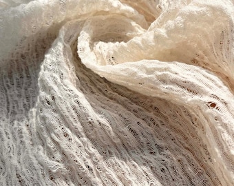 Può includere: Primo piano di un tessuto color crema, a trama larga. Il tessuto ha una trama aperta e strutturata con un aspetto leggermente stropicciato. La luce crea ombre e riflessi, enfatizzando la consistenza e il drappeggio del materiale. L'effetto complessivo è morbido e arioso.