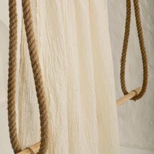 May include: A cream-colored, textured fabric hanging from a wooden rack suspended by thick, brown rope. The rack has two horizontal wooden bars and is supported by the rope looped over the bars. The fabric has a soft, wrinkled appearance.