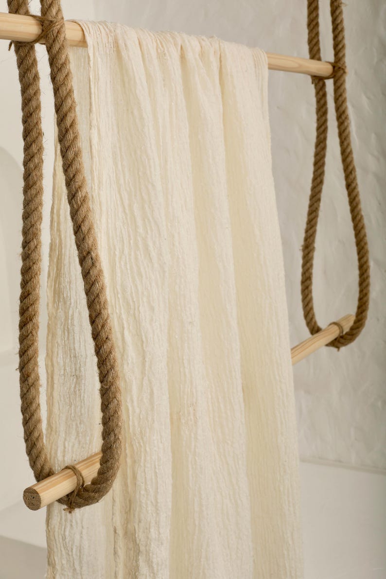 May include: A cream-colored, textured fabric hanging from a wooden rack suspended by thick, brown rope. The rack has two horizontal wooden bars and is supported by the rope looped over the bars. The fabric has a soft, wrinkled appearance.