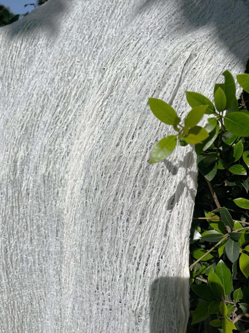 May include: Close-up of a white, textured fabric. The fabric has a loose weave, creating a semi-transparent effect. The texture appears soft and slightly crinkled. Green leaves are visible in the background, adding a natural contrast to the white material.