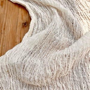 May include: Off-white, textured fabric with a crinkled appearance. The material is draped, creating soft folds and shadows. The fabric's weave is visible, adding to its unique texture. The background is a wooden surface.