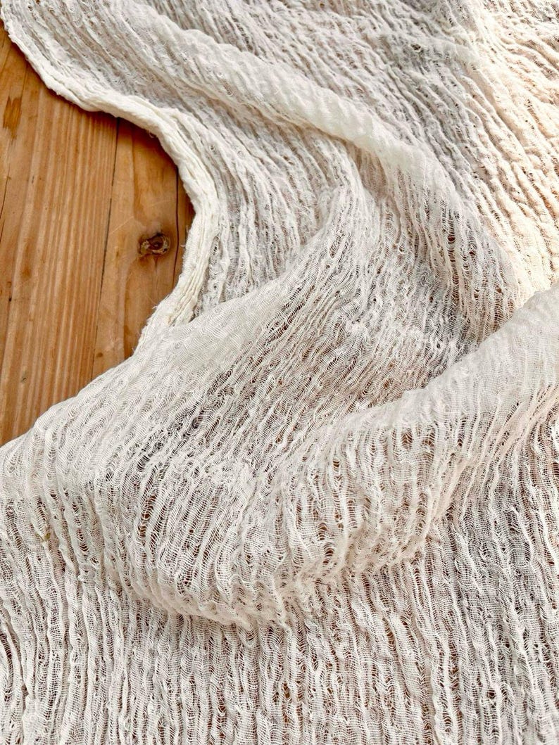 May include: Off-white, textured fabric with a crinkled appearance. The material is draped, creating soft folds and shadows. The fabric's weave is visible, adding to its unique texture. The background is a wooden surface.