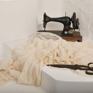 Pu&ograve; includere: Una macchina da cucire vintage con una base in legno si trova su una superficie bianca, parzialmente coperta da un tessuto color crema. Sulla superficie si trova anche un paio di forbici arrugginite.