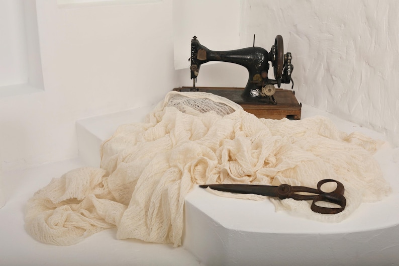 Pu&ograve; includere: Una macchina da cucire vintage con una base in legno si trova su una superficie bianca, parzialmente coperta da un tessuto color crema. Sulla superficie si trova anche un paio di forbici arrugginite.