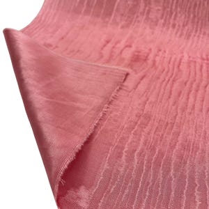 May include: A close-up of a folded piece of pink fabric. The fabric has a textured, ribbed appearance with vertical lines and a shiny finish. The edge of the fabric is frayed, revealing the raw edge of the material.