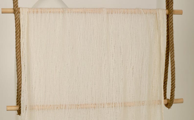 Pu&ograve; includere: Uno sfondo in tessuto di uncinetto bianco appeso a un'asta di legno con una corda marrone. Il tessuto ha un aspetto testurizzato e intrecciato.