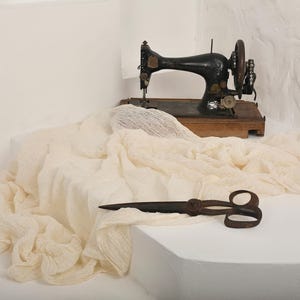 May include: An antique black sewing machine with a wooden base sits atop a white surface. A cream-colored fabric is draped around the machine, and a pair of vintage scissors rests in the foreground. The scene evokes a sense of vintage craftsmanship.