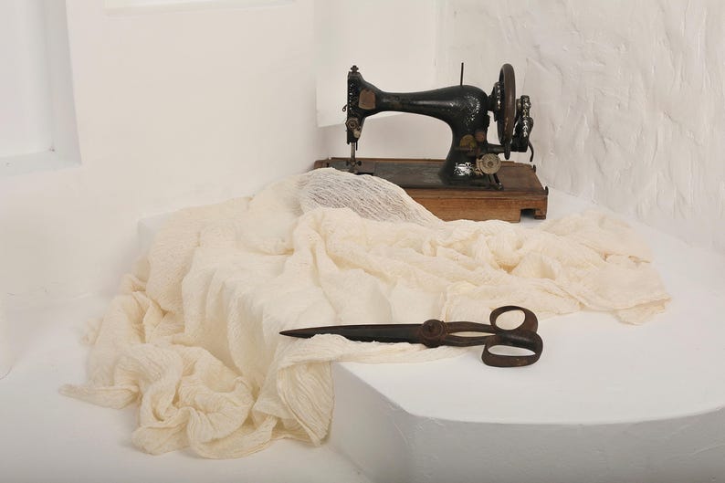 May include: An antique black sewing machine with a wooden base sits atop a white surface. A cream-colored fabric is draped around the machine, and a pair of vintage scissors rests in the foreground. The scene evokes a sense of vintage craftsmanship.
