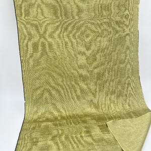 May include: A length of olive green fabric with a wood grain pattern. The fabric is folded over at the bottom right corner, revealing a lighter green underside. The edges are finished with a black border.