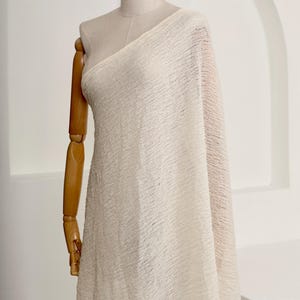 May include: Off-white fabric draped over a dress form. The fabric has a textured, crinkled appearance. The dress form is made of wood and has articulated arms. The background is a plain white wall with an arched architectural detail.