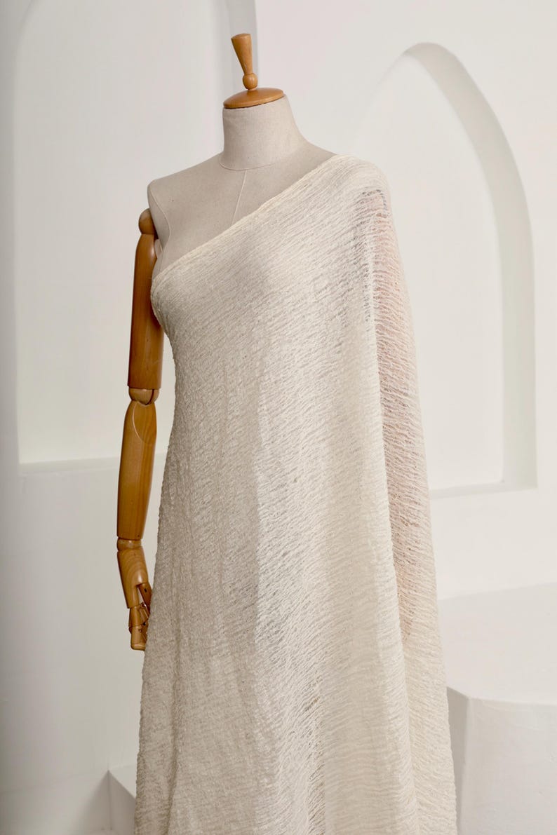 May include: Off-white fabric draped over a dress form. The fabric has a textured, crinkled appearance. The dress form is made of wood and has articulated arms. The background is a plain white wall with an arched architectural detail.