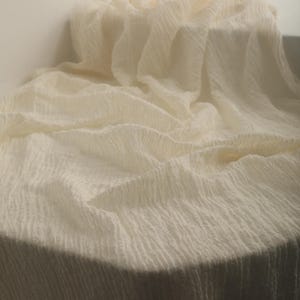 May include: Off-white, textured fabric draped over a round surface. The fabric has a crinkled appearance, with vertical lines creating a subtle pattern. The lighting highlights the folds and texture of the material.