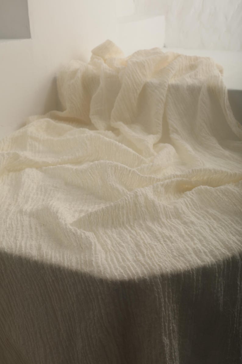 May include: Off-white, textured fabric draped over a round surface. The fabric has a crinkled appearance, with vertical lines creating a subtle pattern. The lighting highlights the folds and texture of the material.
