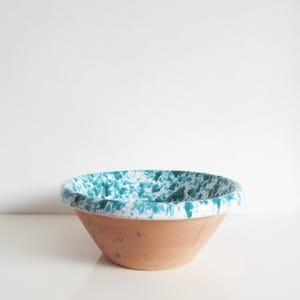Op de afbeelding: Een terracotta kom met een witte rand en een turquoise gespikkeld patroon aan de binnenkant. De kom heeft een brede opening en een afgeronde basis. De kom wordt waarschijnlijk gebruikt voor het serveren of mengen van voedsel.