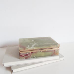 Può includere: Una scatola rettangolare realizzata in pietra lucida verde e bianca con venature rosse prominenti. Il coperchio e la base sono uniti da un sottile bordo metallico color oro che circonda il perimetro, creando una netta separazione tra le parti.