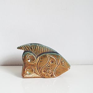 Op de afbeelding: Een keramische vissculptuur in bruin- en tinten. De vis heeft een textuur met uitgesneden details, waaronder cirkels en lijnen. De sculptuur wordt tentoongesteld op een wit oppervlak.