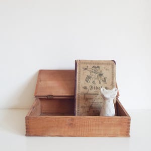 Op de afbeelding: Een rechthoekige houten doos met een scharnierend deksel en vingerlasverbindingen op de hoeken. In de doos ligt een verouderd, beige boek met de titel Fibel en een vintage illustratie van twee kinderen, naast een klein, wit katvormig beeldje.