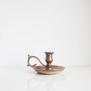 May include: A vintage brass candlestick holder with a handle. The candle holder has a round base and a cup for the candle. The handle is curved and attached to the side of the base. The brass has a warm, aged patina.