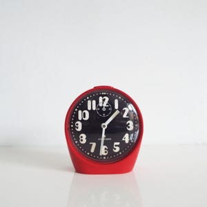 May include: A red, retro-style alarm clock with a black face and white numerals. The clock has a curved, rounded design and the brand name "Junghans" is visible on the face.