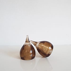 Op de afbeelding: Twee bruine glazen flessenstoppers met een druppelvorm en lange, dunne tuiten. Eén stopper staat rechtop, de andere ligt op zijn kant. Het glas is doorschijnend en laat licht door. Geschikt voor wijnflessen.