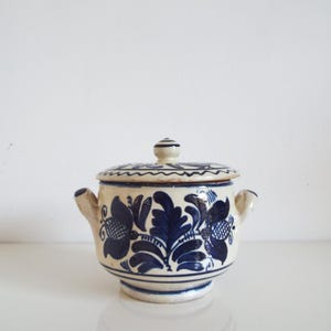 May include: A ceramic lidded bowl with a cream base and blue floral designs. The bowl has two small handles and a knob on the lid. The floral pattern includes stylized flowers and leaves. The bowl is likely handmade.