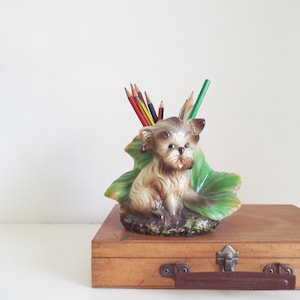 Puede incluir: Un portalápices con forma de perro de cerámica con una base de hoja verde. El perro es de color marrón claro y crema, con la boca ligeramente abierta. Varios lápices de colores están en el soporte. El soporte está sobre una caja de madera.