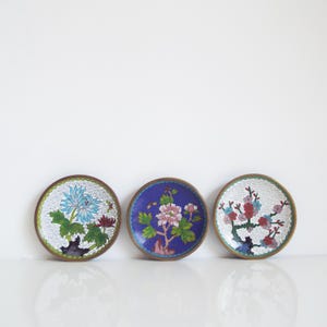 以下が含まれることがあります： 花柄のデザインが施された、3つの小さくて丸い装飾皿。それぞれ異なる配色で、青い花、青い背景にピンクの花、白い背景に赤とピンクの花が描かれています。すべて金色の縁取りが施されています。
