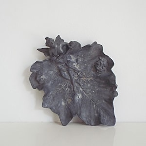 Può includere: Un piatto decorativo a forma di foglia, grigio scuro, con una figura di fata scolpita. La fata è appollaiata sulla foglia, che presenta venature dettagliate. Il piatto ha una finitura opaca ed è probabilmente realizzato in metallo o materiale simile.