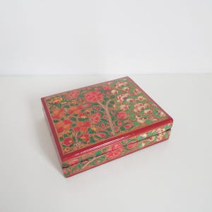 May include: A rectangular, red wooden box with a floral design. The box features a detailed pattern of red, orange, and green flowers and leaves. The edges are trimmed with gold. The box is closed and sits on a white surface.