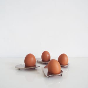 Può includere: Quattro uova di gallina marroni riposano in portauova individuali in metallo color argento. Ogni portauovo presenta una base piatta e arrotondata con piccoli piedini, progettata per sostenere un singolo uovo in posizione verticale.