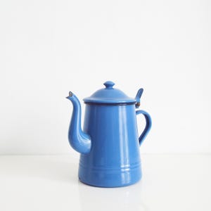 May include: A bright blue enamel coffee pot with a matching lid, a curved spout, and a side handle. The pot features a smooth, glossy finish with a dark rim around the top edge and a small metal hinge connecting the lid to the main body.