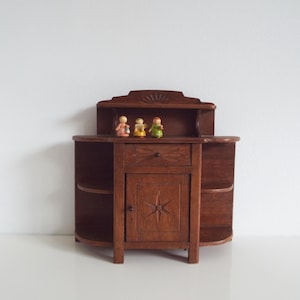 以下が含まれることがあります： 濃い茶色の仕上げが施された、小さなアンティークの木製キャビネット。引き出し、星型のデザインのキャビネットドア、湾曲したサイドシェルフがあります。3つの小さな置物が上に立っています。
