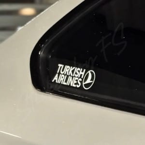 Könnte beinhalten: Weißer Aufkleber mit dem Logo "Turkish Airlines", einschließlich des Namens der Fluggesellschaft und eines stilisierten Globus. Der Aufkleber befindet sich auf einer schwarzen Oberfläche, wahrscheinlich einer Autoscheibe, mit einer weißen Karosserie.